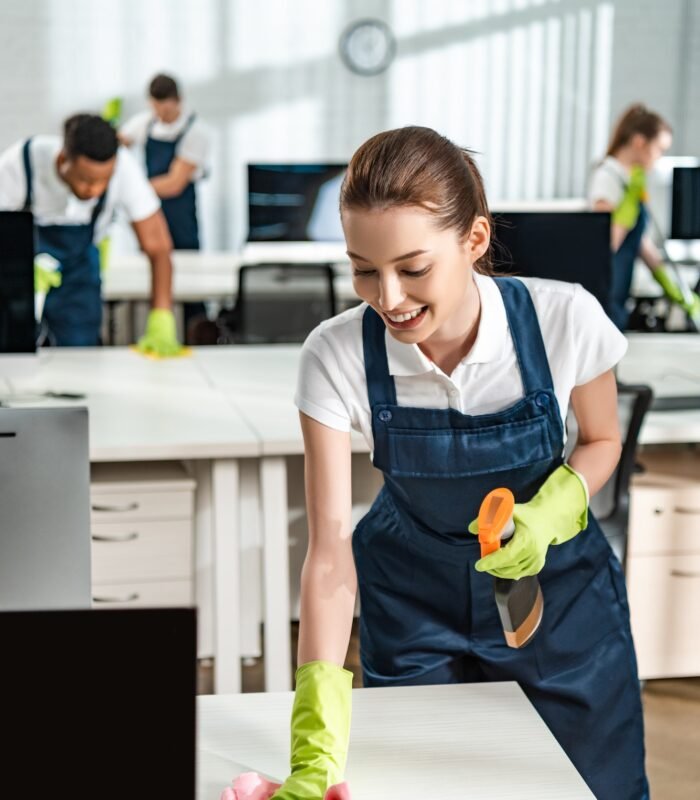 cheerful cleaner in overalls cleaning office desk with rag