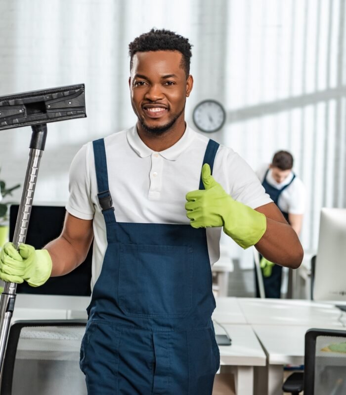 smiling african american cleaner holding vacuum cleaner brush and showing thumb up near team of